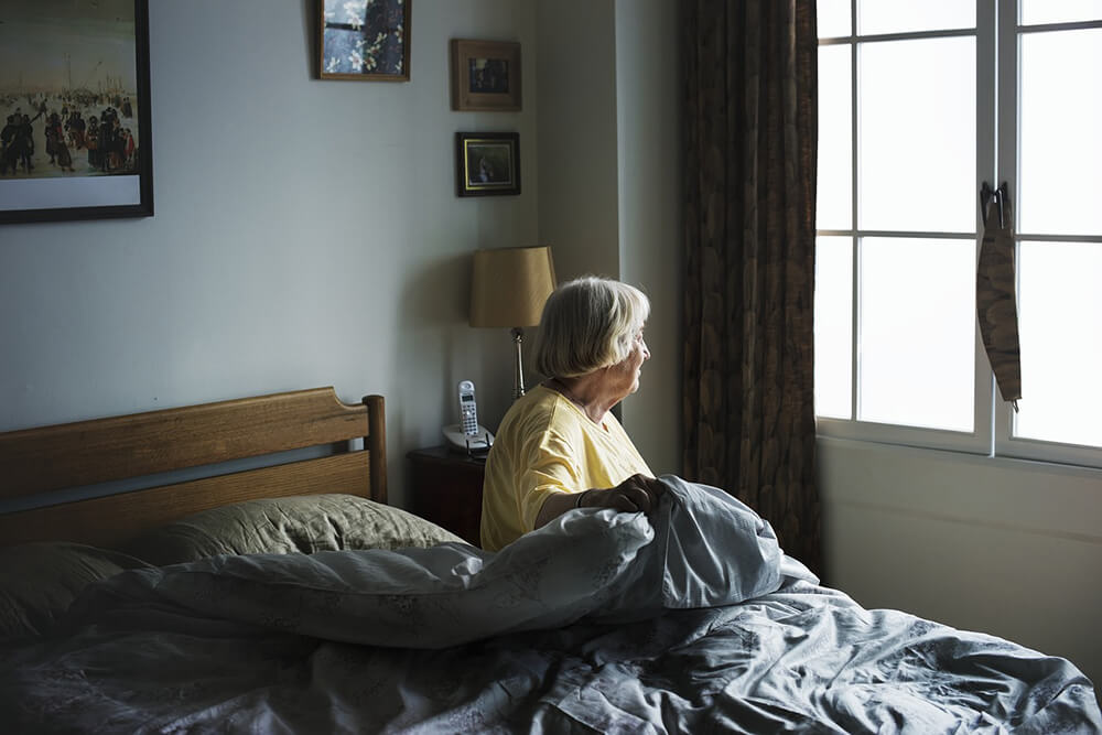 Elderly woman sitting on bed looking out window 